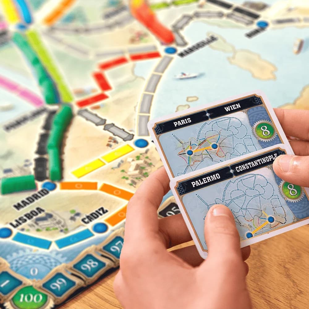 Een hand met twee bestemmingskaarten boven het bord van Ticket to Ride